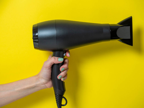 secador de pelo negro en la mano de una mujer sobre un fondo amarillo. Cuidado del cabello Producto - Foto, Imagen