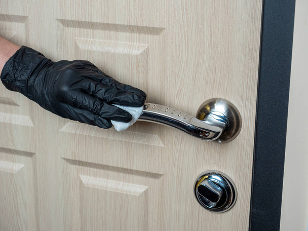 de hand van een man in een zwarte latex handschoen veegt het handvat van de voordeur af met een doek. Desinfectie van het interieur door virussen en microben - Foto, afbeelding