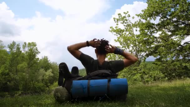 Explora el mundo y disfruta de la vida. Joven hombre caucásico guapo con rastas está sentado en la naturaleza apoyándose en una gran mochila turística y estirando los brazos hacia arriba. Viajero solo, corto metraje de 4k. - Metraje, vídeo