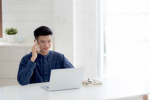 Jonge Aziatisch man praten telefoon en werk vanuit huis met laptop computer, freelance met behulp van notebook naar internet online en spreken op smartphone, huishoudelijk leven, zakelijke en communicatie concept. - Foto, afbeelding