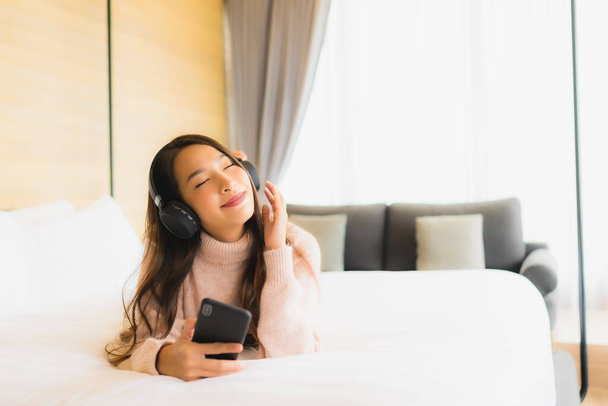 Asyalı güzel bir kadının portresi. Yatak odasının içinde müzik dinlemek için kulaklıklı cep telefonu kullanıyor. - Fotoğraf, Görsel