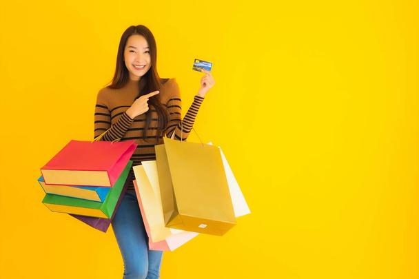 肖像美しい若いアジアの女性は黄色の隔離された背景にカラフルなショッピングバッグで買い物のためのクレジットカードを使用 - 写真・画像