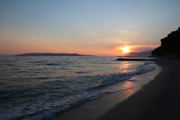 Coucher de soleil panoramique sur la plage au bord de la mer croate. Paysage marin soir mer et soleil jaune à l'horizon, soleil éblouissant à la surface de la mer, roches et mousse de mer un soir d'été - Photo, image