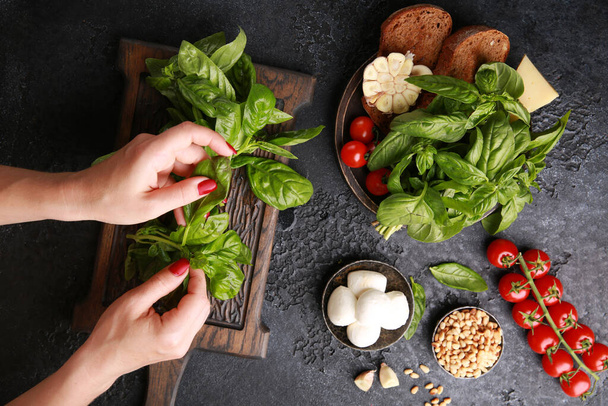 Italian cuisine. Preparation, cooking process with hands. Ingredients: basil, mozzarella, tomatoes, garlic, bruschetta, pine nuts on a dark table. Background image, copy space. Flatlay, top view - 写真・画像
