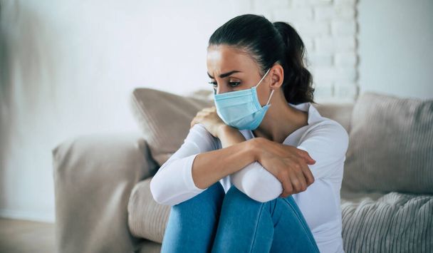 Young woman in deep depression is sitting on the couch in safety medical mask during coronavirus quarantine - Foto, Imagem