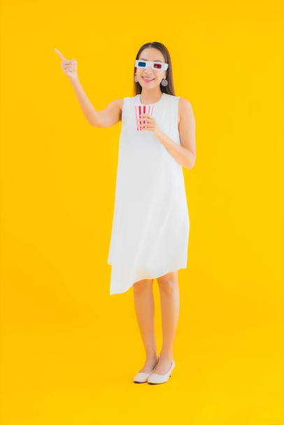Portret mooie jonge aziatische vrouw met popcorn en 3d bril op geel geïsoleerde achtergrond - Foto, afbeelding