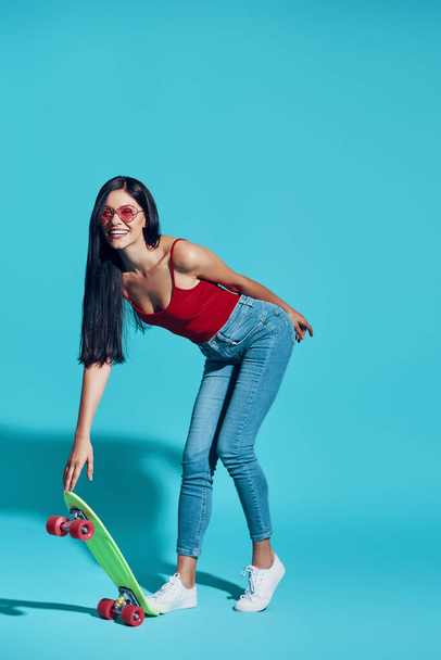 Full length of attractive young woman looking at camera and smiling while skateboarding against blue background - Foto, Imagem