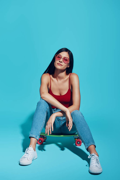 Attractive young woman looking at camera while sitting on skateboard against blue background - Foto, Imagem