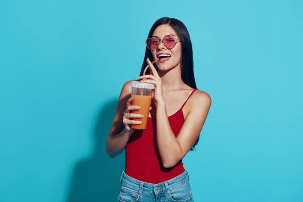 Attractive young woman holding cup with drink and smiling while standing against blue background - Photo, Image