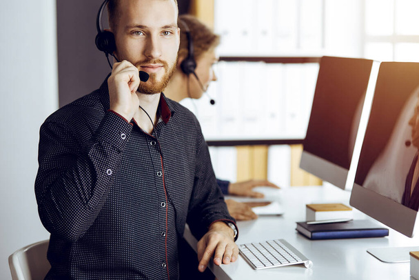 Bearded businessman talking by headset near his female colleague while sitting in modern sunny office. Diverse people group in call center - Foto, Imagem