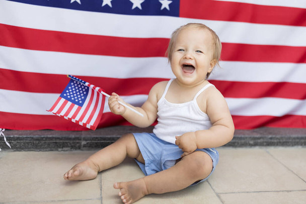 Patriotic holiday. Cute caucasian baby ten-eleven months old with blonde hair and blue eyes with American flag at home. USA celebrate independence day 4th of July. - Foto, Bild