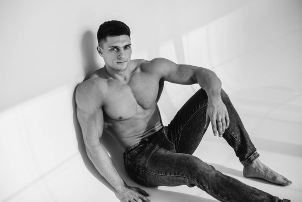 A young sexy athlete with perfect abs is sitting in the studio topless in jeans. Healthy lifestyle, proper nutrition, training programs and nutrition for weight loss. Black and white. - Fotoğraf, Görsel