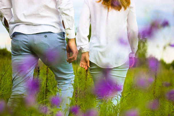 Glückliches verliebtes Paar beim Spazierengehen auf einer Frühlings- und Sommerwiese. Mann von hinten, Frau in weißen Hemden, Jeans. Eine wunderschöne Naturlandschaft, violett blühende Blumen. Ein Date, Romantik, Beziehungskonzept.  - Foto, Bild