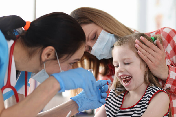 Dokter in beschermende medische masker nemen buccaal uitstrijkje van klein meisje met wattenstaafje - Foto, afbeelding