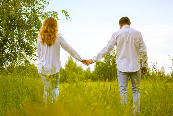 Happy couple in love walks in a spring, summer meadow, holding the hands. Back view husband, wife in white shirts. A beautiful natural green landscape. Newlyweds. A date, romance, relationship concept - Photo, Image