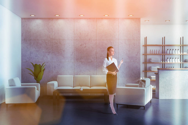 Attractive woman holding black folder with membership contract, waiting for clients in reception area of modern luxury spa wellness center to sign in documents. Towels, soaps and perfumes on shelves - Фото, зображення