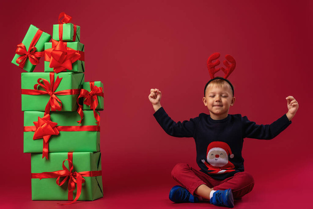 little boy in Santa Claus hat sits next to pyramid of Christmas gift boxes on red background in studio. child is happy to relax and meditate and receives a lot of gifts for Christmas. The advertiser. - Fotó, kép