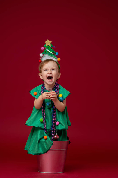little girl in a Christmas tree costume stands in a bucket on a red background. The child is very upset, crying and screaming, very unhappy. - Zdjęcie, obraz
