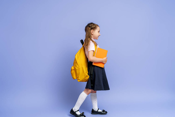 Cute happy kid in uniform. raises his leg high and runs on purple background. child with backpack. little girl is ready for school. Dynamic images that go back to conceptual school. holidays begin. - Foto, Imagen
