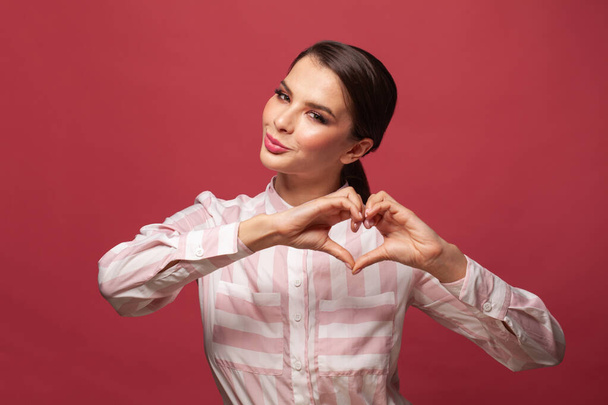 Happy young woman showing heart with hands. Portrait of beautiful smiling brunette woman wearing shirt and showing heart with hands. - Foto, afbeelding