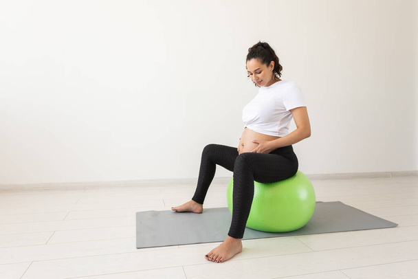 Молодая беременная женщина делает расслабляющие упражнения, используя фитнес-мяч, сидя на коврике. Copyspace. - Фото, изображение