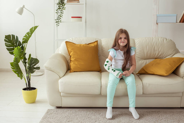 Niño con un yeso en una muñeca o brazo roto sonriendo y divirtiéndose en un sofá. Actitud positiva, recuperación y concepto de niño. - Foto, imagen