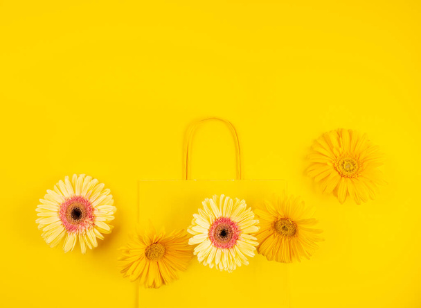 Κίτρινο επίπεδη χάρτινη τσάντα αγορών με λουλούδια μαργαρίτα ζέρμπερα σε μονόχρωμο φόντο - Φωτογραφία, εικόνα