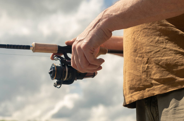 Fisherman hands holding spinning reel rod or spoon bait and fishing close up. Summer sky with clouds on background - Fotografie, Obrázek