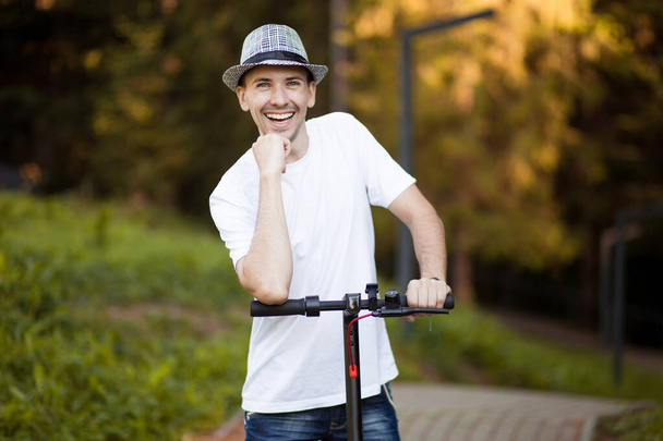 Portré fiatal jóképű férfi fehér ingben, napszemüveg és kalap készen áll, hogy lovagolni az elektromos robogó kerékpár a város utcáján napsütéses nyáron. Ökológiai közlekedési koncepció. Elektromos szállítás. - Fotó, kép