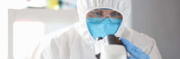 Врач в защитном медицинском костюме, маске и очках смотрит через микроскоп - Фото, изображение