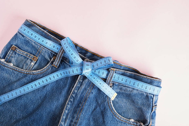 blue measuring tape is threaded into jeans instead of a belt, blue jeans on a pink background, copy space, weight loss and weight control concept, measuring body volume - Fotoğraf, Görsel