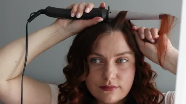 Belle jeune femme fait des boucles coiffure avec un fer à friser à la maison. Femme souriante coiffure ondulée boucles - Séquence, vidéo