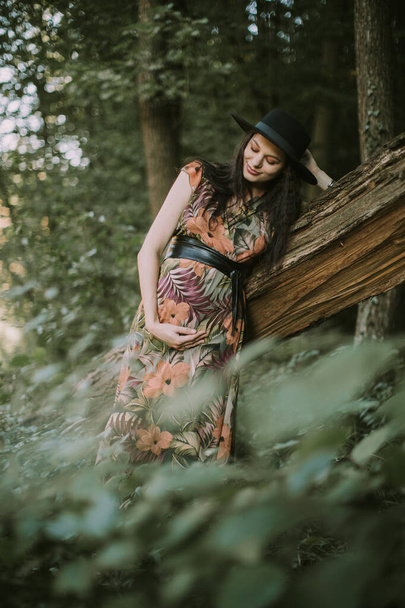 Portrait of a happy and pretty pregnant woman looking at her belly, leaning on the tree outdoors - Φωτογραφία, εικόνα