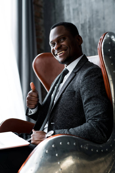 Przystojny, uśmiechnięty afrykański biznesmen, siedzący na krześle i podnoszący kciuk. - Zdjęcie, obraz