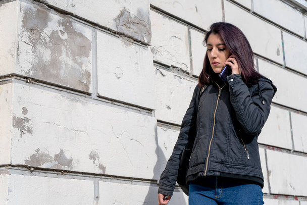 American shot of Young Latina woman walking and talking with a worried expression on cell phone in an old city street on a cold autumn morning. Telecommunications, cell phone concept. - Foto, Imagen