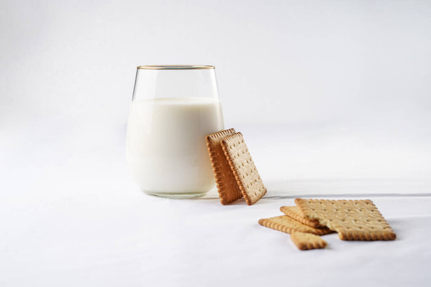 Стакан молока на белом скатерти и свежеиспеченные печенья, опирающиеся на стекло - Фото, изображение