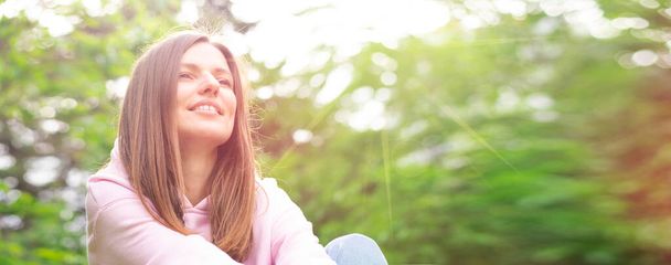 ピンク色のセーターを着た緑の芝生の上に座っている若い陽気な女性の肖像画(パーカー)公園の日没時の人の全体像. - 写真・画像