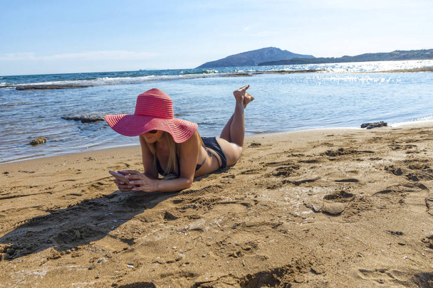 nő fekete bikini és piros kalap feküdt trópusi strandon nézi a mobiltelefon pihentető nap alatt - Fotó, kép