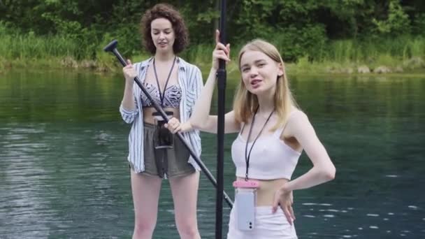 Deux jeunes femmes heureuses debout sur des planches dans la rivière et regardant dans la caméra - Séquence, vidéo