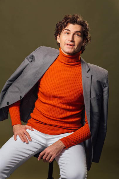 Portrait of a tall handsome man dressed in orange turtleneck, grey jacket and white jeans posing on the green background - Фото, изображение