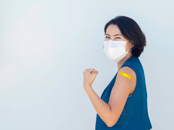 Impfungen, Verband am Arm geimpfter Menschen. Glückliche asiatische Geschäftsfrau mit blauer Blazerweste, Gesichtsmaske mit gelbem Verband und selbstbewusster Geste nach der Impfung. - Foto, Bild