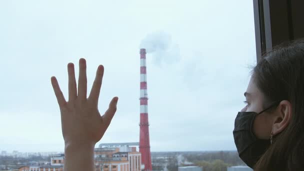 Una chica con una máscara negra mira por la ventana, poniendo su mano sobre el cristal. Contaminación ambiental - Imágenes, Vídeo