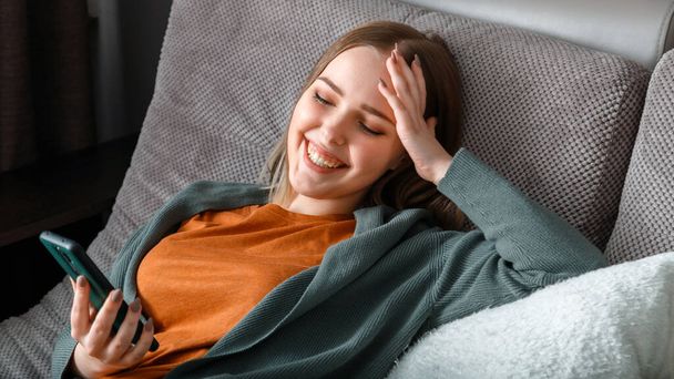 Happy smiling teenage girl laughing texting on phone surfing Internet or chatting using smartphone lying on couch. Young woman use smartphone for leisure enjoyment laughs. Long web banner - Фото, зображення
