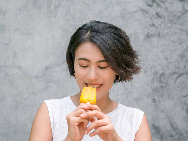 Woman eating popsicles. Happy beautiful Asian woman short hair wearing casual white sleeveless shirt holding yellow popsicle, outdoors. Smiling female enjoying ice lolly in summer. - 写真・画像