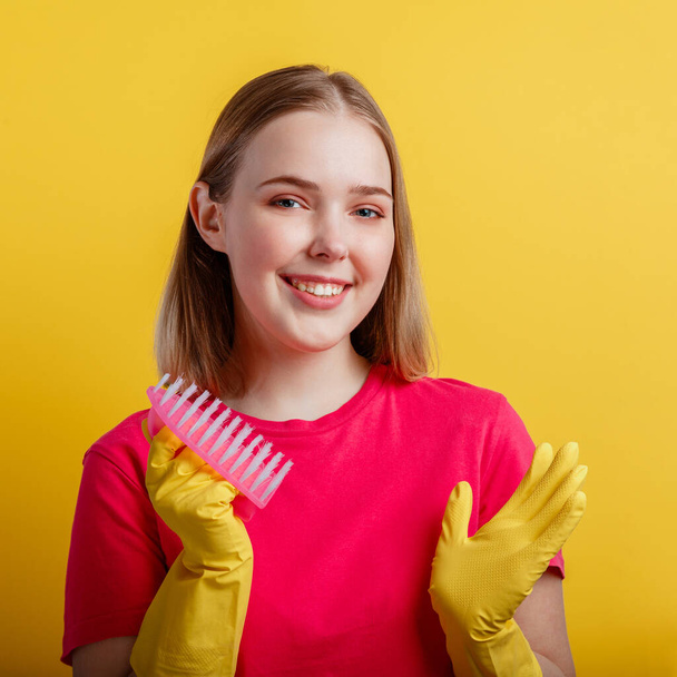 ラバー手袋のクリーニングブラシと女性の肖像画。若いブロンドの幸せな笑顔の女性は黄色の色の背景に隔離された家庭用品で家を掃除する準備ができています。クローズアップ広場 - 写真・画像