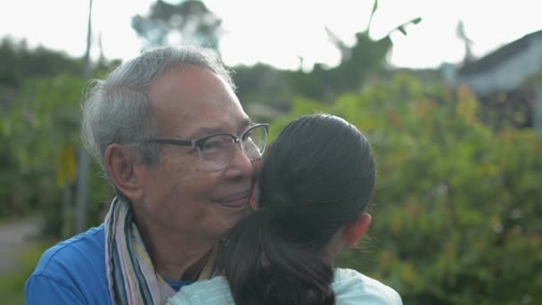 Gelukkige Aziatische senior grootvader in bril omarmt zijn jonge kleindochter die terug te komen om hem te bezoeken op het platteland thuis. Uitdrukking van liefde in twee generaties. Samenhorigheid. Landelijk gebied. - Video