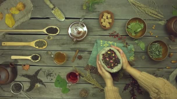 Czerwona herbata z płatków róż na starym drewnianym stole. Leżeć płasko. Martwa natura z różnymi rodzajami herbaty: czarny, zielony, mate, hibiskus, kwiatowy, owocowy, ziołowy. Kobiece dłonie położyły na stole torebkę herbaty z hibiskusem. Zwolniony ruch 2x. - Materiał filmowy, wideo