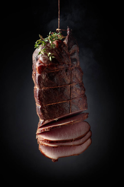 Sliced smoked gammon with thyme branches on a black background. Natural product from an organic farm, produced by traditional methods. - Photo, Image