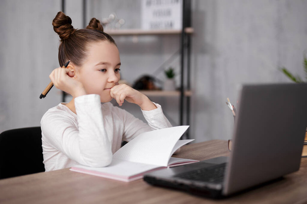 school, homework and distance education concept - portrait of cute thoughtful girl doing homework and using laptop for studying online at home - Photo, Image