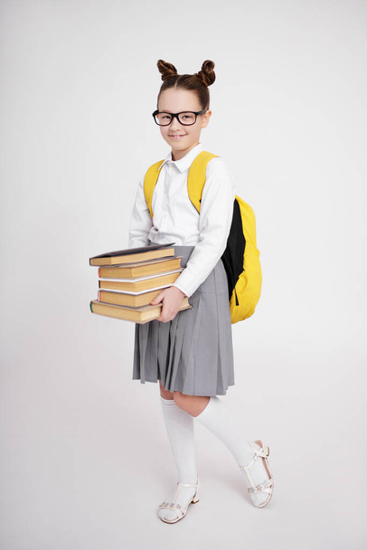 concetto di istruzione - ritratto a figura intera di ragazza carina in uniforme scolastica e occhiali con zaino in possesso di libri su sfondo bianco - Foto, immagini